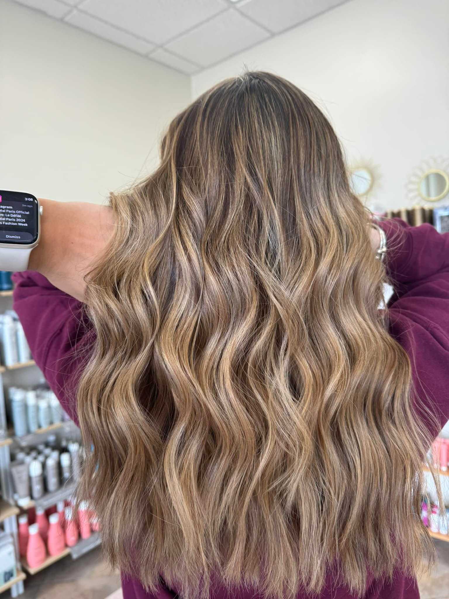 Woman with wavy, highlighted hair in a salon, wearing a smartwatch and maroon top.