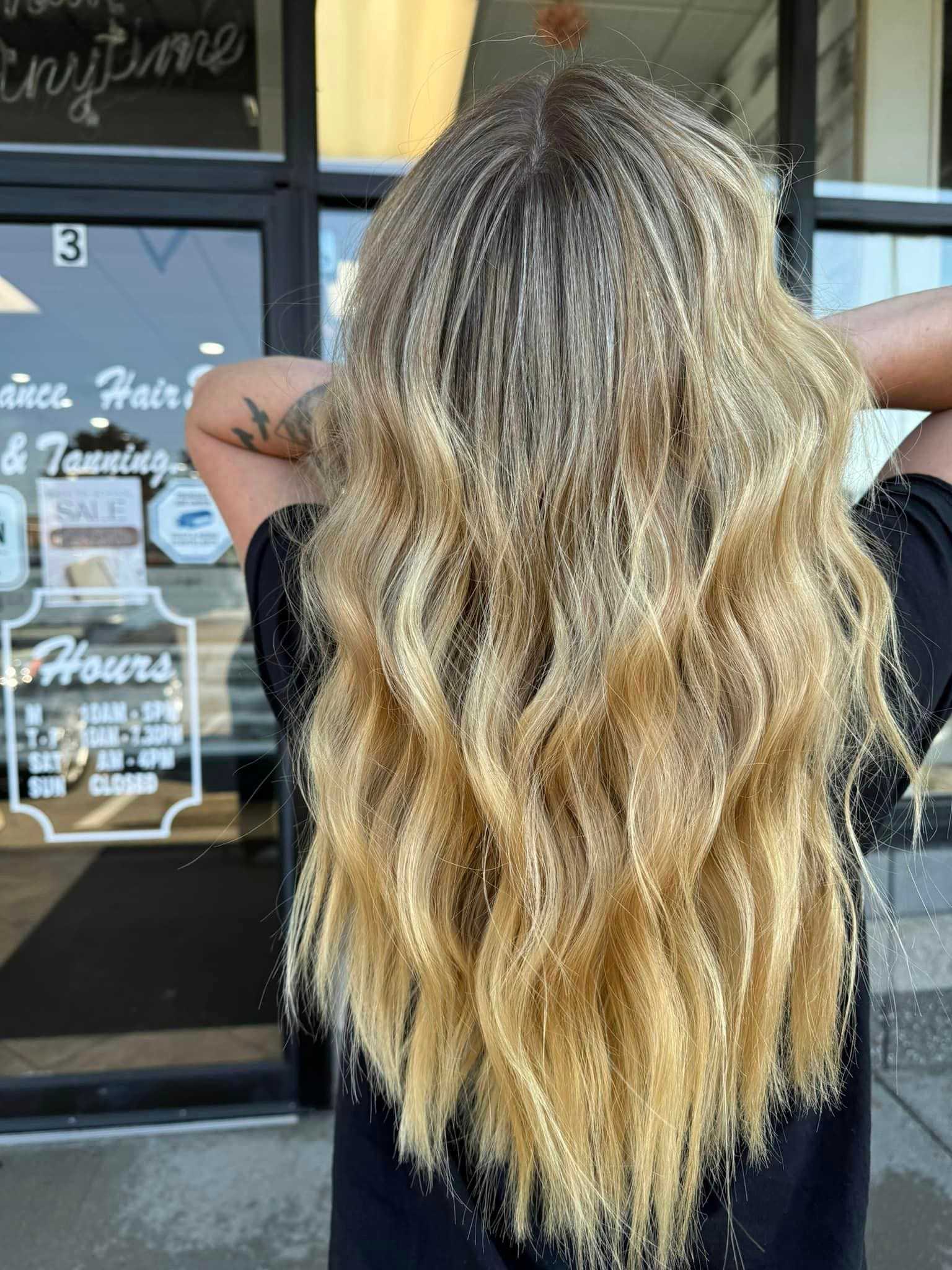 Woman with long, wavy blonde hair standing in front of a hair salon entrance.