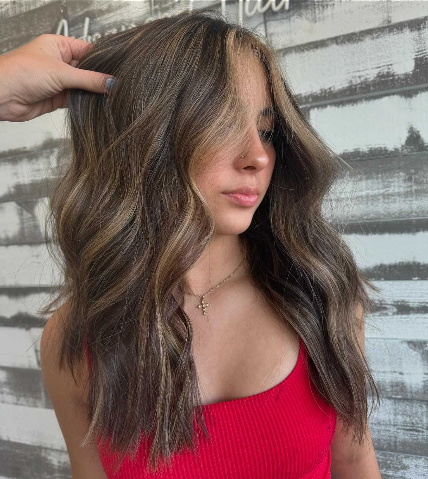 Woman with wavy brown hair and blonde highlights, wearing a red top, posing in front of a textured wall.
